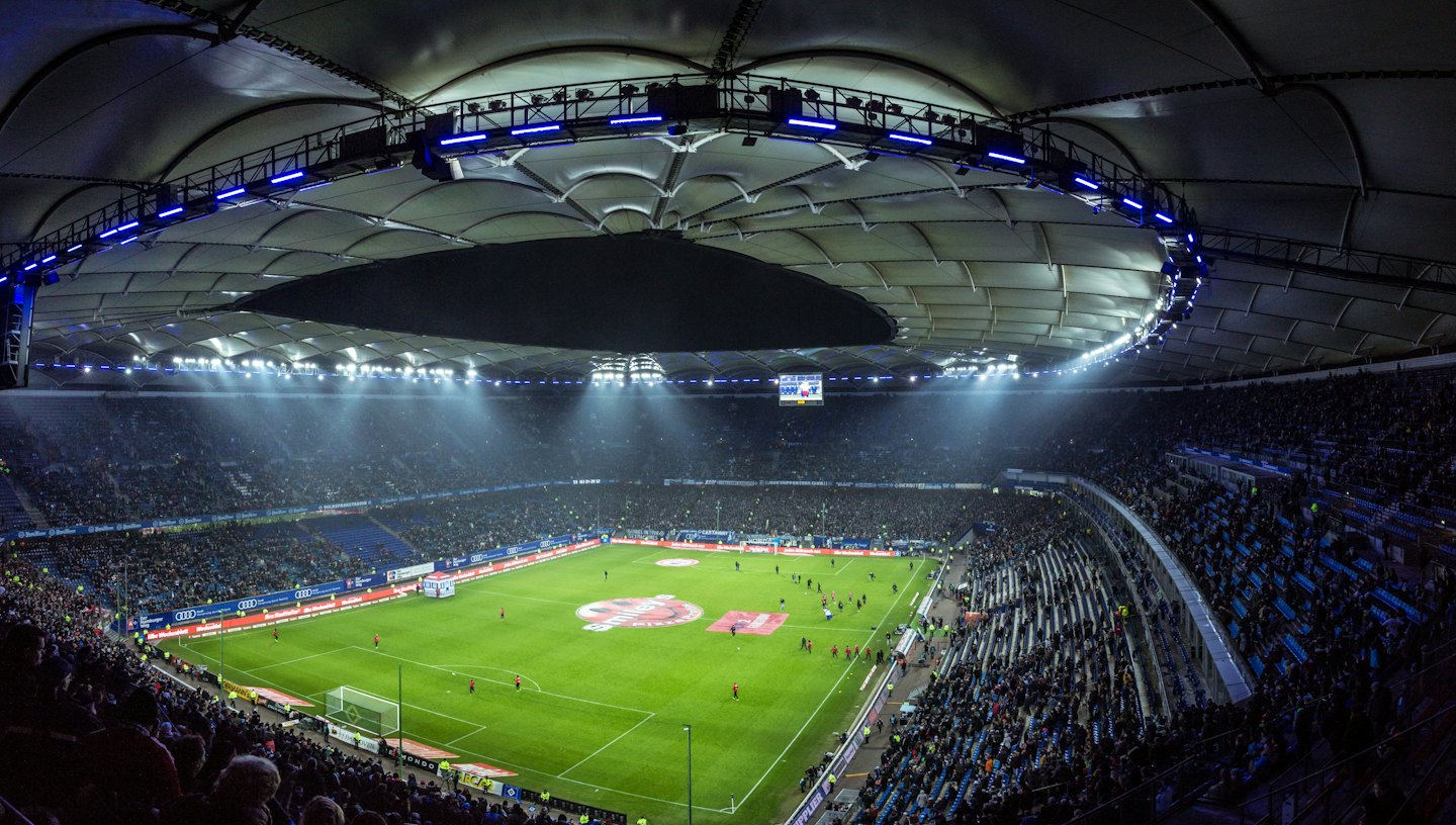 Modern football stadium filled with fans at night under bright lights.